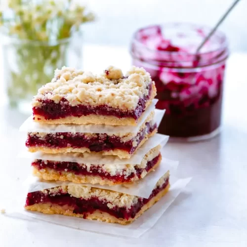 Blueberry crumb bars with homemade jam stacked on a white surface with a jar of berry jam in the background